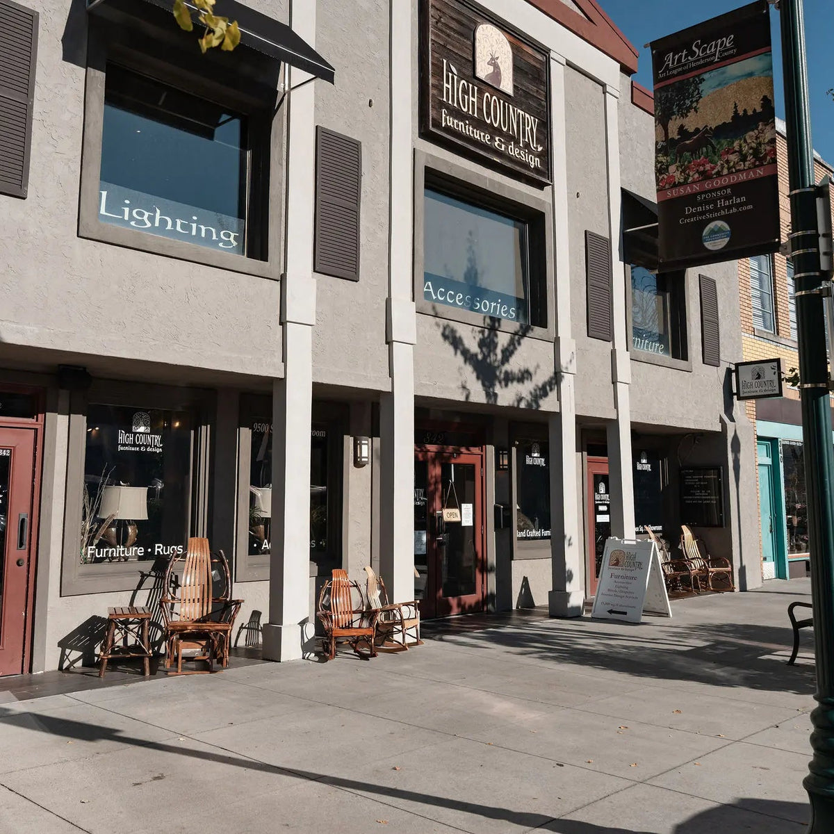 A street-facing storefront scene with multiple commercial buildings, windows, awnings, outdoor chairs, a sidewalk, and a green lamppost under a blue sky.
